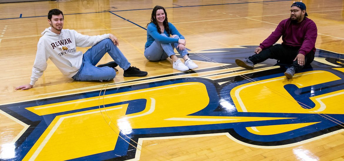 Rowan students sit on a gymnasium floor at a community college, showing the school's logo.