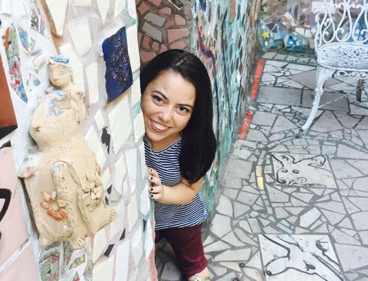 Mackenzie Trush smiles from behind a wall filled with mosaic art