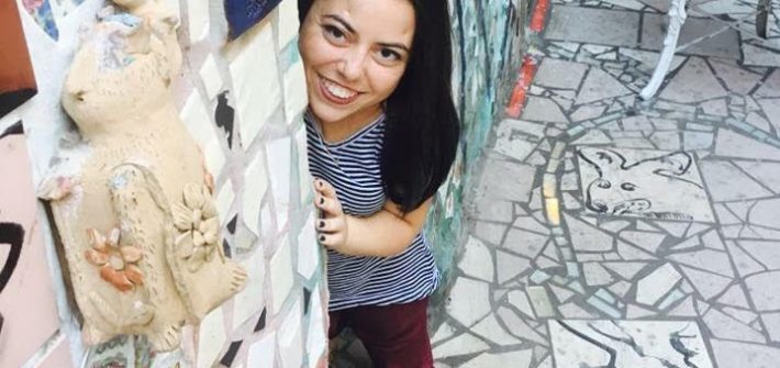 Mackenzie Trush smiles from behind a wall filled with mosaic art