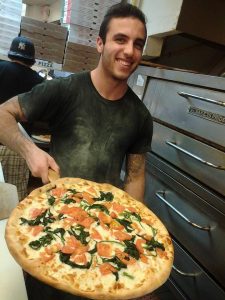Kyle holds a pizza coming out of a pizza oven, while smiling