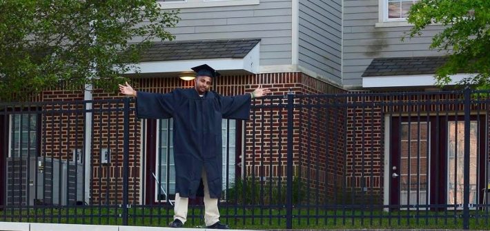 Sha stands in his cap and gown with arms outstretched on top of a Rowan sign