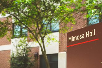 brown sign for Mimosa Hall, in front of building with blooming green trees