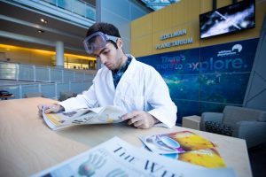 student in lab coat inside science all reading the newspaper