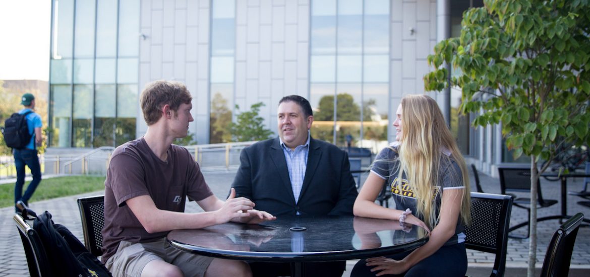 Eric Liguori sitting outside with two students outside the Rohrer College of Business