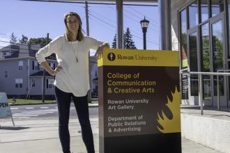 student outside communications building at Rowan University, hand on hip and other leaning on the building sign