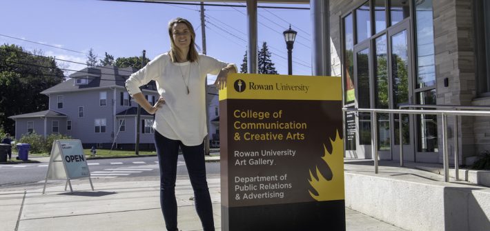 student outside communications building at Rowan University, hand on hip and other leaning on the building sign