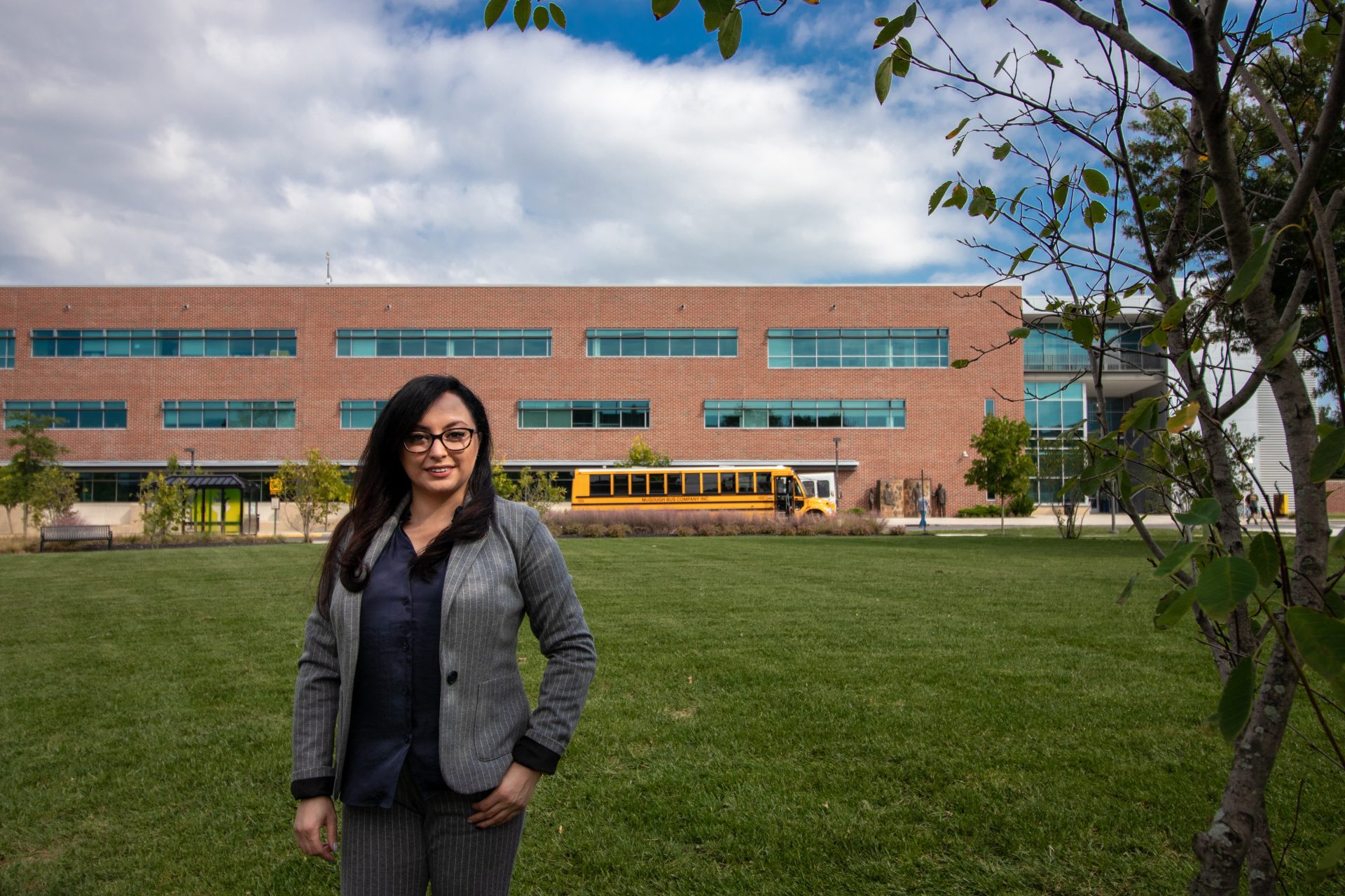 business woman standing on grass with brick building and yellow school bus in the distance
