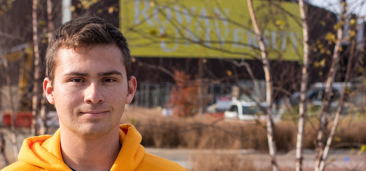 Young, white male student with yellow Rowan University standing in foreground with brick building blurred out in background