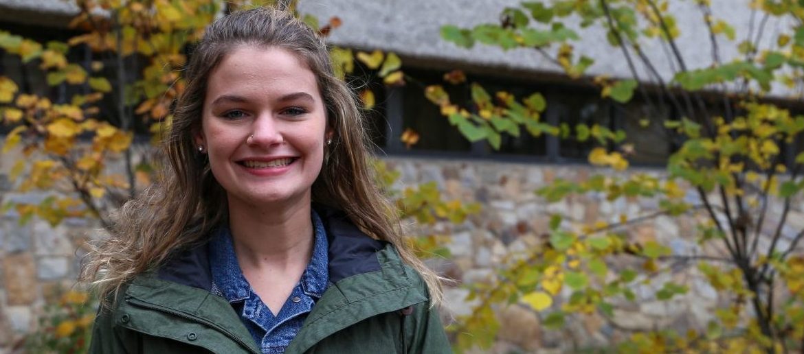Sarah stands in front of autumn trees outside of Robinson Hall, smiling and wearing a green jacket