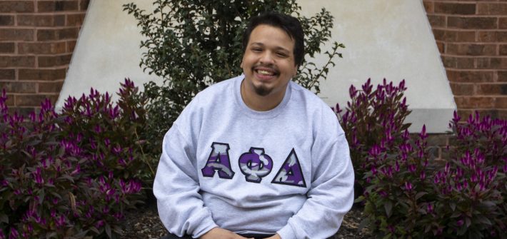 Victor sits on the brick area outside the student center while wearing a Greek Alpha Phi Delta sweatshirt
