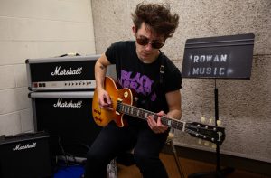 Luke sitting playing guitar in a practice room in Wilson Hall with his amps 