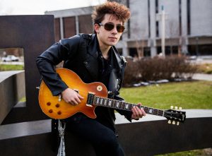 Luke sitting on statue outside Robinson circle holding his guitar in a leather jacket and sunglasses 