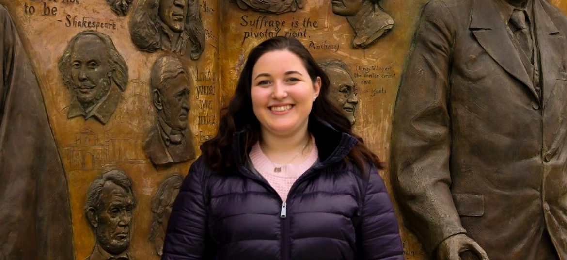 Rebecca Shnier stands outside of James Hall at Rowan University, in front of a bronze artwork that says Knowledge is Power