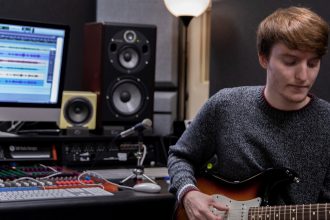 Young male student playing electric guitar inside a music production student while sitting down in a chair