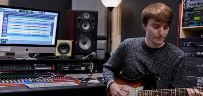 Young male student playing electric guitar inside a music production student while sitting down in a chair