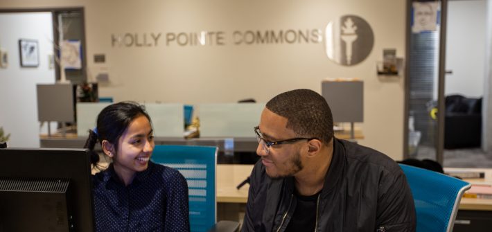 two students inside Holly Pointe behind front desk smiling at each other