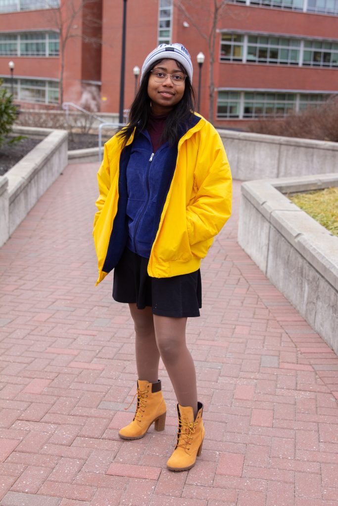 A rowan student in a beanie, yellow jacket and tan boots stands by Savitz hall