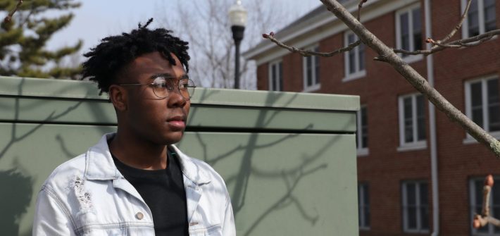 Jelani wears a ripped white denim jacket over a black t-shirt, standing in front of a pale green metal wall near dorms at Rowan University