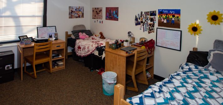 Wide view of students' dorm with two beds, two desks, and a window with its blinds down