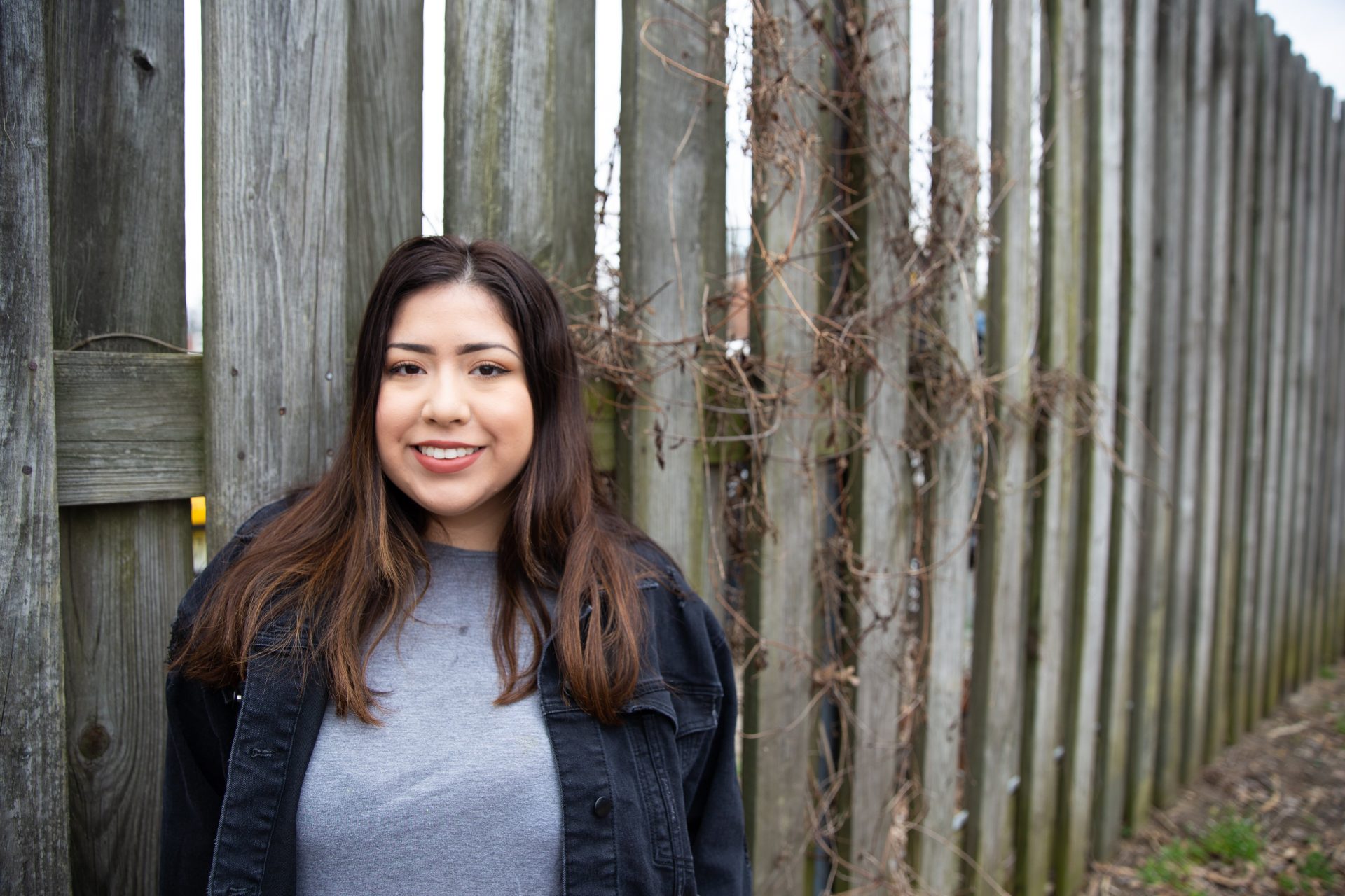  A portrait of Jenn Cruz posing in front of a wooden fence on campus.