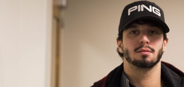 Robert stands in his doorway of Holly Pointe at Rowan University wearing a black jacket and staring at the camera