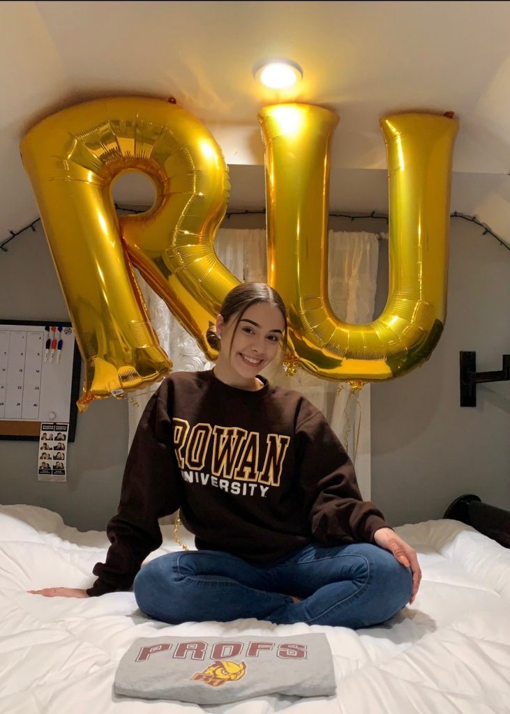 Arianna in her room wearing a rowan sweater next to large gold "RU" balloons