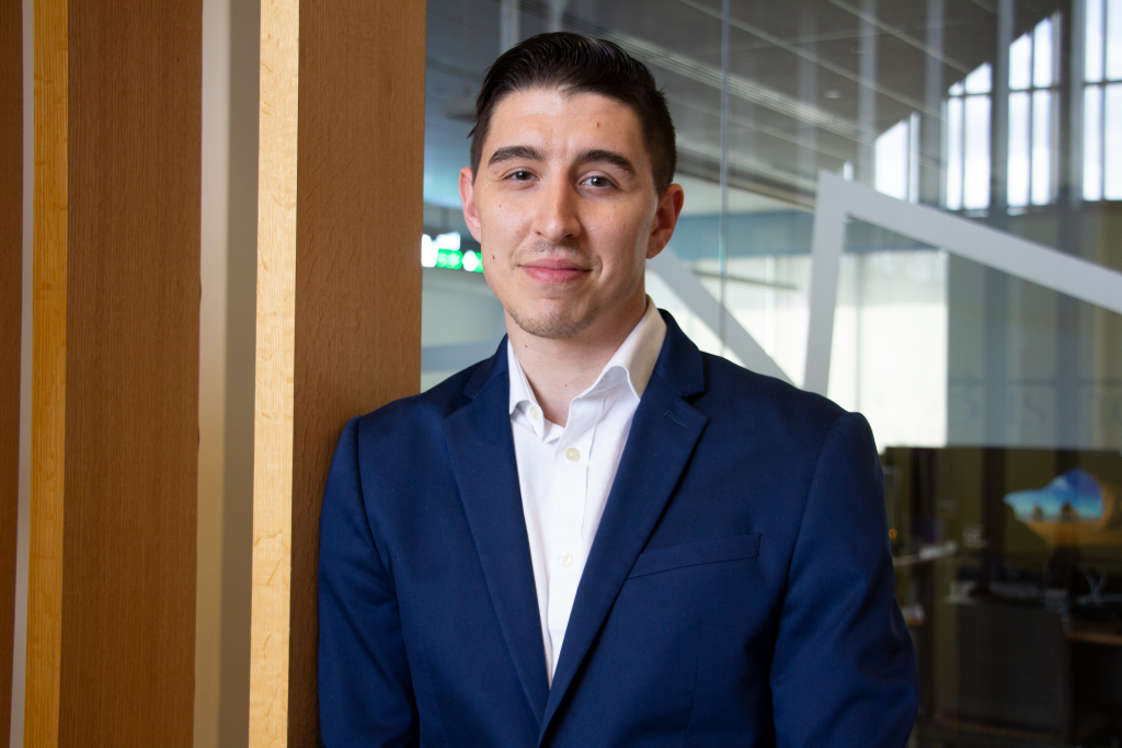 Zac, wearing a blue blazer leans against a wall in Business Hall