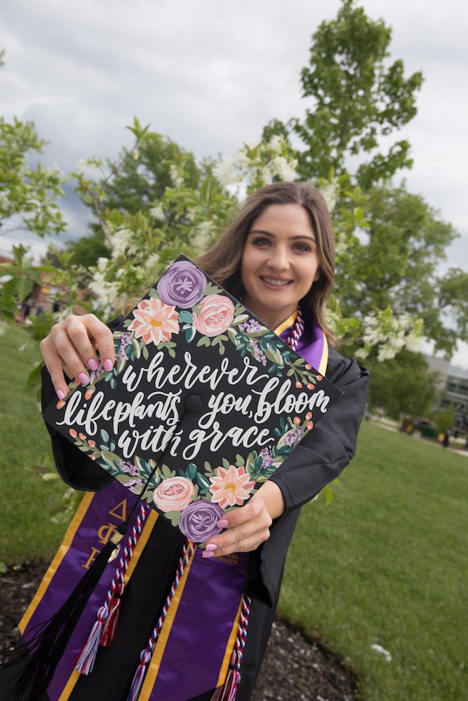Brianna in her graduation gown holding up her decorated cap that says "wherever life plants you, bloom with grace"