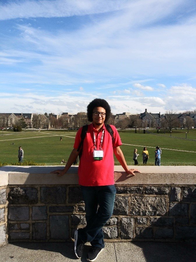Student in red shirt and jeans standing against waist-high stone wall with field and trees in the background