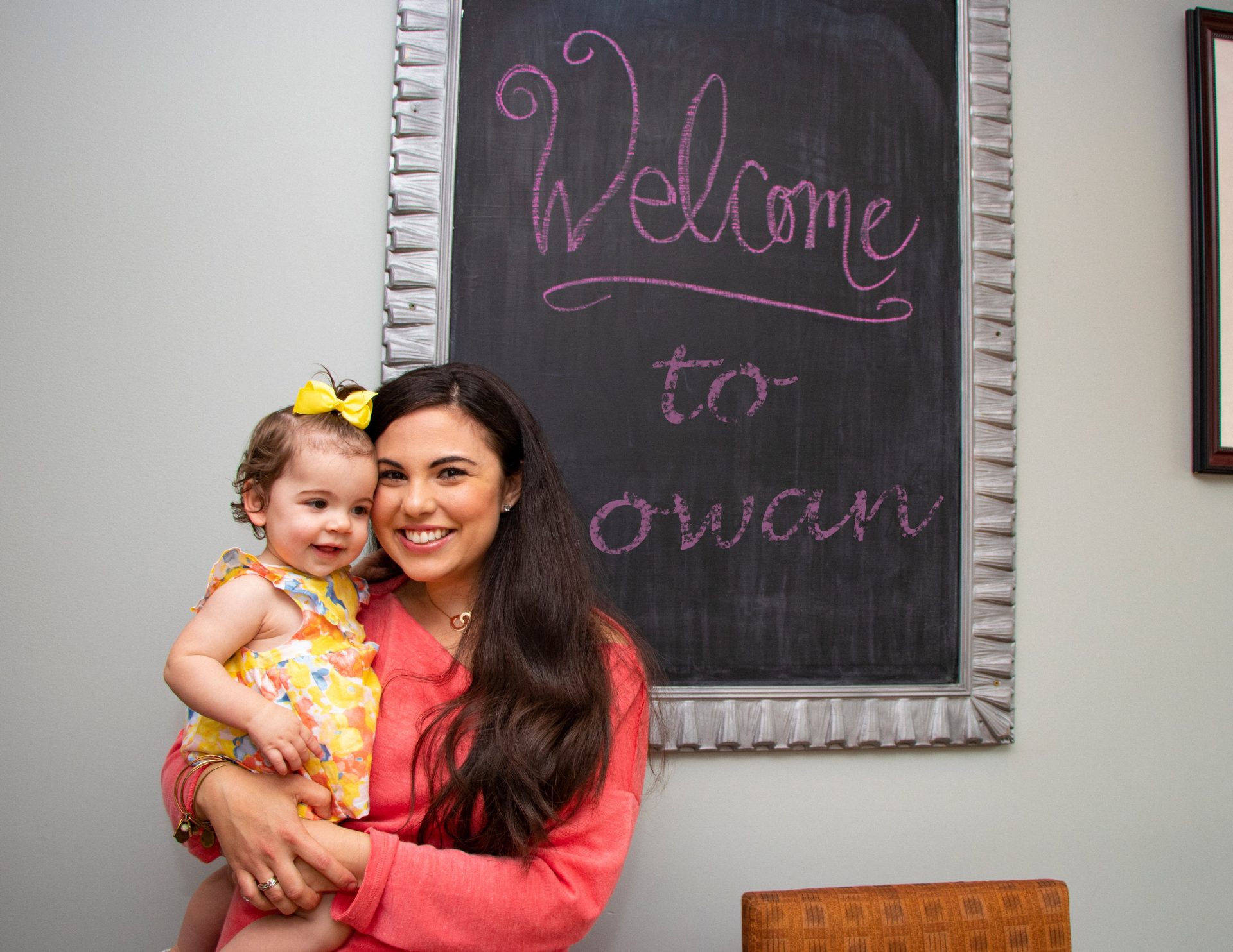 A woman wearing a pink shirt holding a baby girl in front of a decorative chalkboard