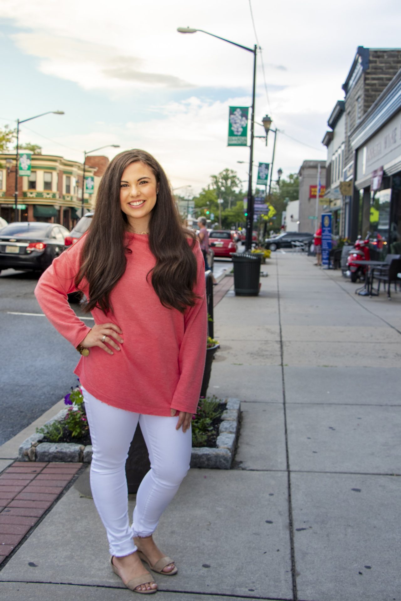 A woman in a pink top and white pants with her hand on her hip posing on a busy town street sidewalk