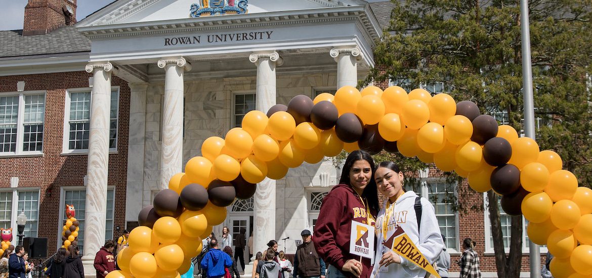 Two young women stand under a brown and gold school colors balloon arch at Rowan University at Accepted Students Day.