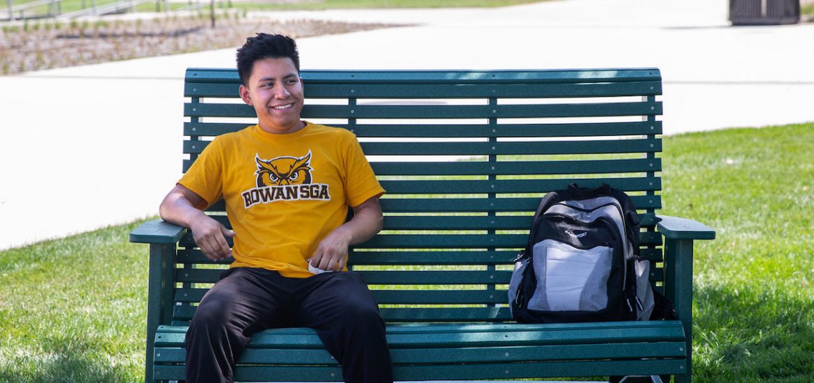 Alexis Benitez smiles, sits on a green bench with his bookbag by his side