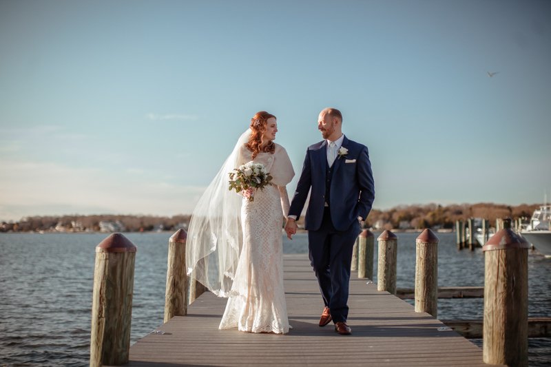 Enchanted Celebrations photo of bride and groom holding hands on a dock with water behind them.