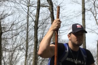 Eddie crouches in the woods, holding a walking stick.