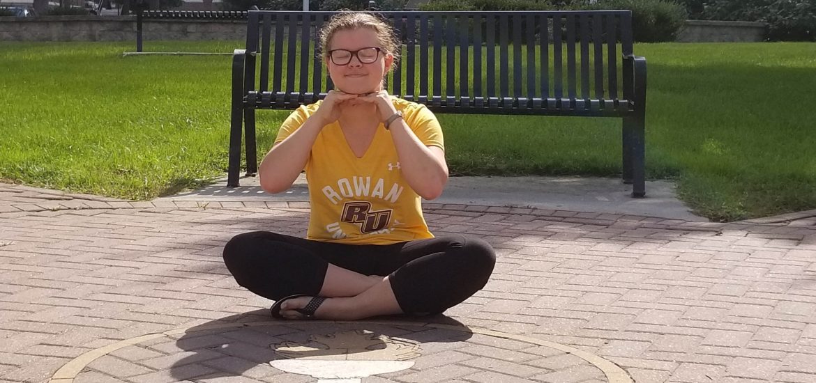 Amanda wears a Rowan shirt and sits on the ground smiling in front of a Rowan logo on the ground.
