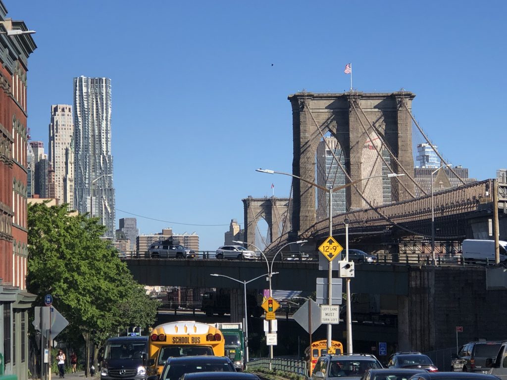the Brooklyn bridge against a clear blue sky, with cars and school buses lined up on the streets below.