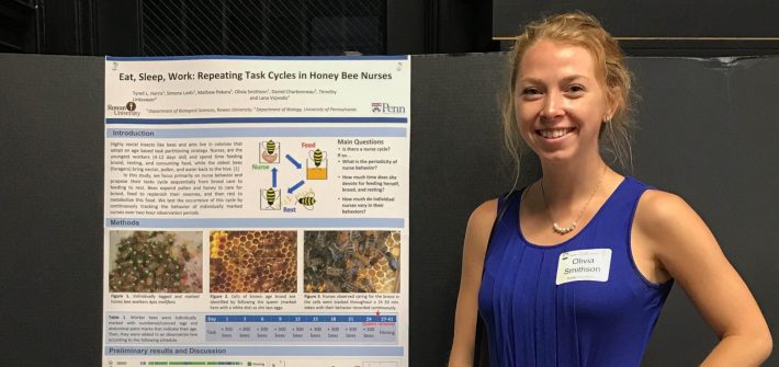 Olivia stands in a blue tank top next to a poster during a presentation.