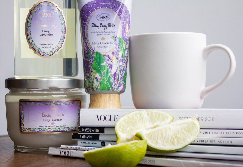 a stack of skincare products from the brand Sabon NYC, next to a coffee mug and slices of lime on a pile of magazines.