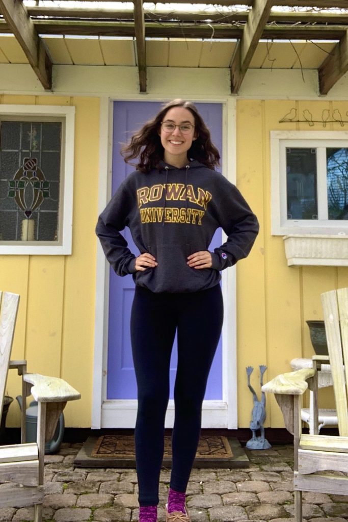 Hannah smiling and posing in front of a yellow shed with a purple door wearing a Rowan University hoodie.