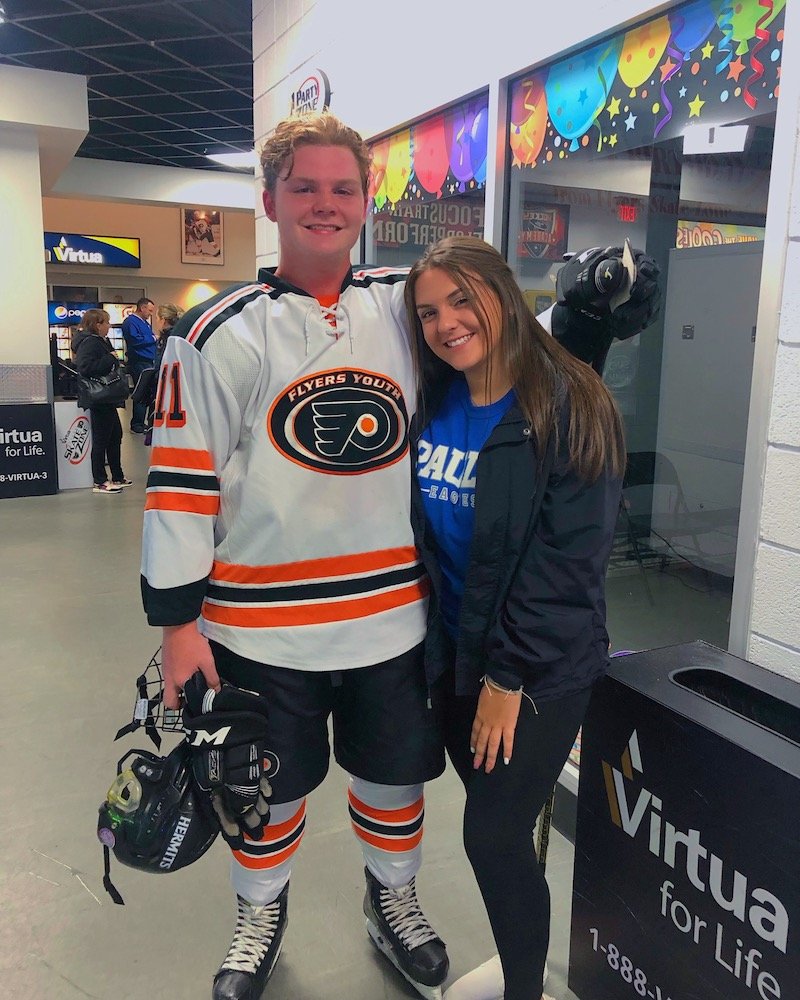 George stands wearing his ice hockey uniform, holding skates, with his arm around a young woman.