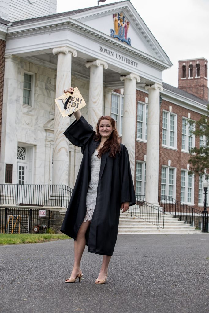 Sarah Transue stands in her graduation gown holding her cap in the air in front of Bunce Hall