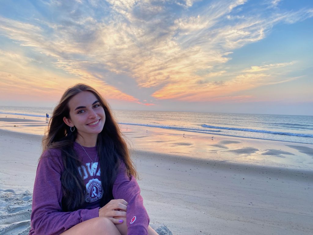 Sydney smiling and sitting on a beach, wearing a Rowan sweatshirt.
