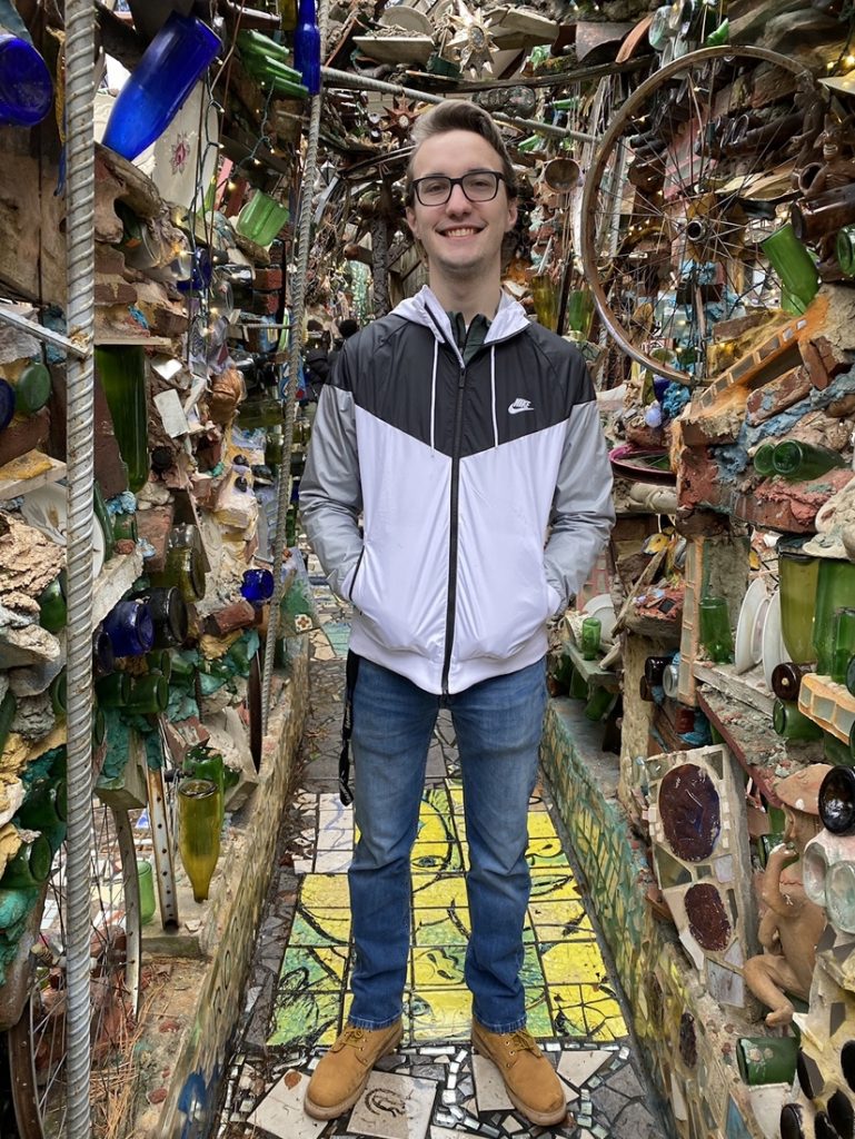 Matthew poses in a hallway with shelves filled with glass objects.