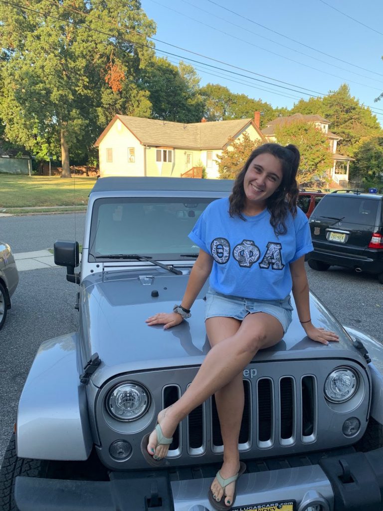 Caroline sits on a Jeep wearing a shirt with her Greek letters