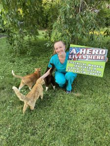 Katelyn posing outside wearing scrubs and holding a healthcare heroes sign alongside her two cats.