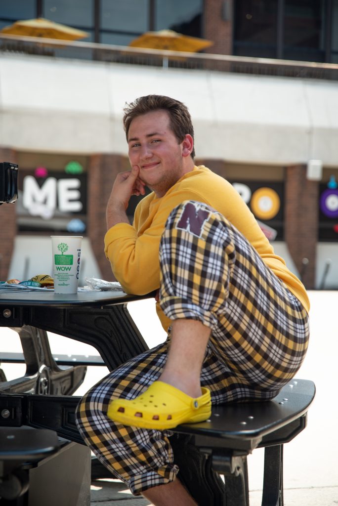 Ben sitting and smiling for a photo outside the student center while wearing yellow crocs and Rowan gear.