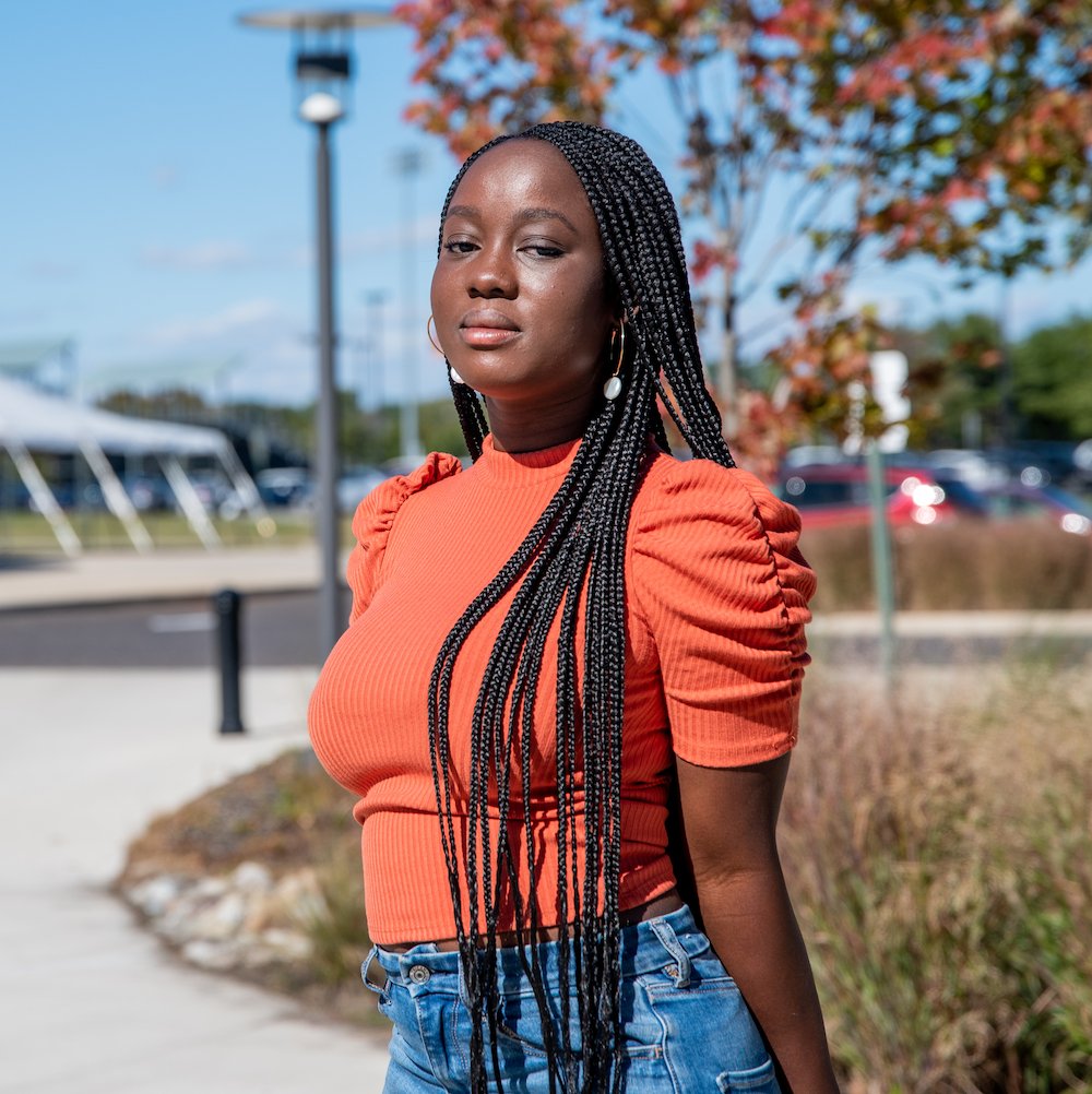 Nafisat Olapade poses in an orange shirt in front of bushes and a parking lot.