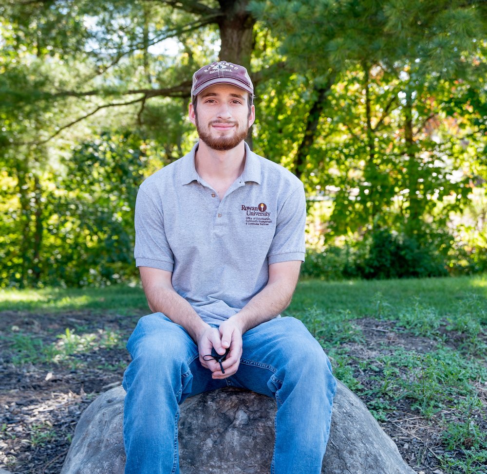 Will sits on a stone in front of trees on campus. 