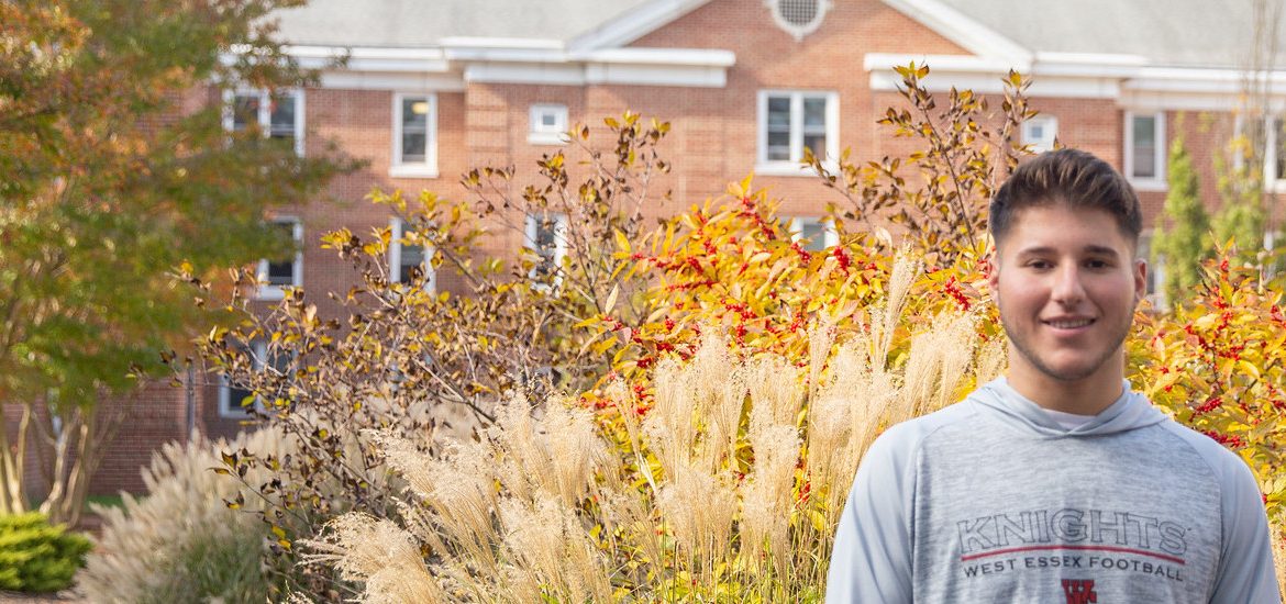 Christopher standing in front of Chestnut Hall. There is green and white shrubbery around him. Trees with green and orange leaves.